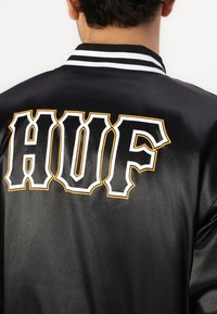Jachetă bomber din satin neagră, cu literă îndrăzneață albă și portocalie „HUF” pe spate. Guler ribbed în dungi negre și albe.