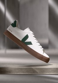 White sneaker with green V logo, featuring a brown rubber sole. Smooth leather upper, lace-up front, and logo on the heel.