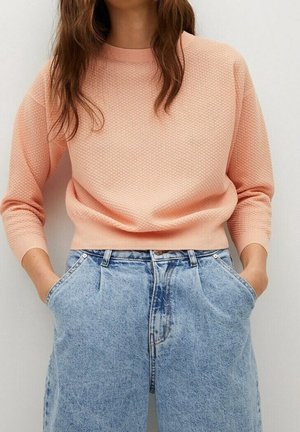 Femme portant un pull rose clair texturé à manches longues, rentré dans un jean en denim bleu clair taille haute, les mains dans les poches.