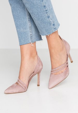 Fødder iført lyserøde ruskind højhælede sko med spidse hæle og tynde krydsremme, kombineret med cropped lyseblå jeans med frynser i kanten.
