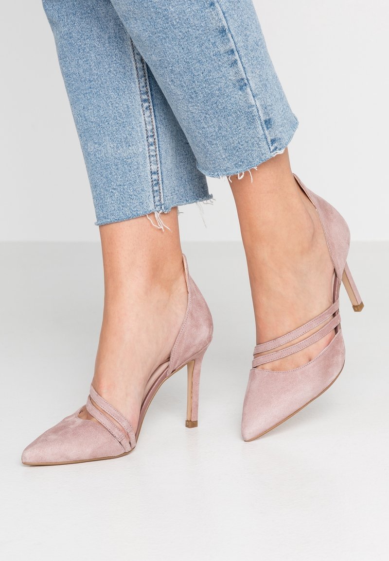 Pieds portant des escarpins en daim rose clair à bout pointu avec de fines brides croisées, assortis à un jean cropped bleu clair à ourlet effiloché.