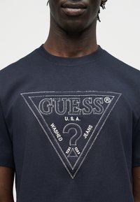 Άνδρας που φοράει μπλε σκούρο μπλουζάκι με λευκό κεντητό λογότυπο Guess και κείμενο σε μορφή τριγώνου στο μπροστινό μέρος.