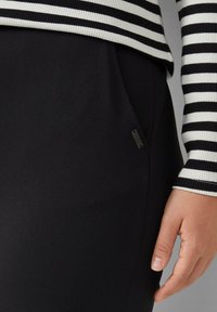 Schwarze Hose mit glatter Textur, ausgestattet mit einer Seitentasche und dezentem Branding. Kombiniert mit einem gestreiften, gerippten Langarmoberteil.