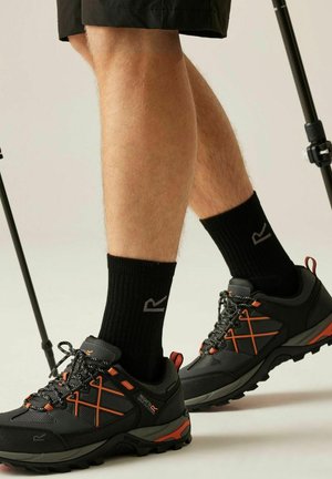 Piernas con zapatos de senderismo negros con detalles naranjas y calcetines negros, de pie con bastones de senderismo contra un fondo liso.