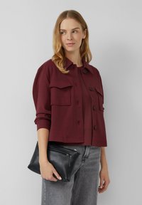 Giacca corta borgogna con due tasche sul petto e chiusura a bottoni, abbinata a un top coordinato e una borsa a tracolla in pelle nera. Jeans grigi visibili.