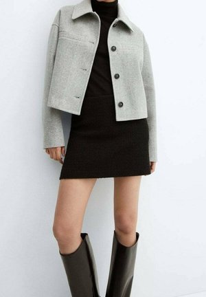 Personne portant une veste courte gris clair avec des boutons, un col roulé noir, une mini-jupe noire et des bottes noires montant au genou sur fond blanc.