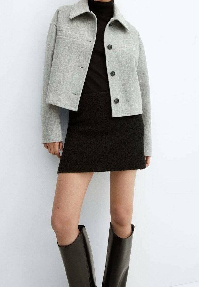 Personne portant une veste courte gris clair avec des boutons, un col roulé noir, une mini-jupe noire et des bottes noires montant au genou sur fond blanc.