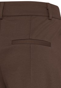 Gros plan sur un pantalon en tissu marron montrant la ceinture arrière, les passants de ceinture et une poche arrière passepoilée unique.