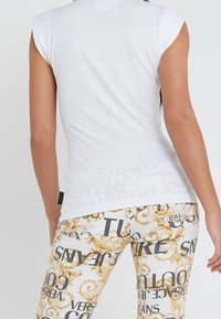 Bílý top bez rukávů s hladkou texturou, zkombinovaný s vzorovanými kalhotami se zlatými akcenty a výrazným černým textem na světlejším pozadí.