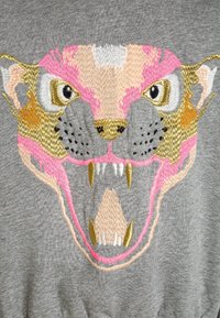 Grijze sweatshirt met een ingewikkeld geborduurd gezicht van een leeuw. Kleuren zijn roze, goud en wit met gedetailleerde gelaatstrekken en tanden.