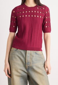 Viininpunainen ribbikuvioinen neulepaita, jossa on lyhyet hihat ja pyöreät aukot hartioilla, yhdistettynä vaaleansinisiin farkkuihin.