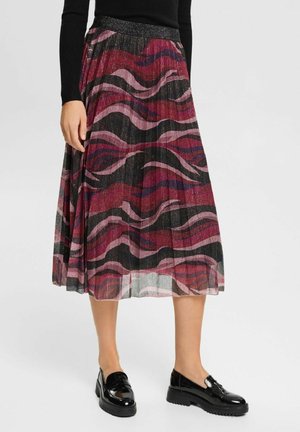 Kvinde iført et plisseret midi-nederdel med rød, pink og sort snoet mønster og skinnende sorte loafers, stående mod en ensfarvet baggrund.
