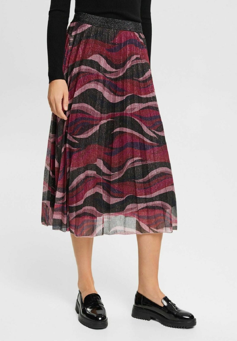 Femme portant une jupe midi plissée avec un motif tourbillonnant rouge, rose et noir et des mocassins noirs brillants, debout devant un fond uni.
