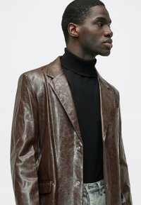 Blazer maro din piele cu finisaj texturat, revere cu decupaj, doi nasturi și un buzunar la piept. Asortat cu un pulover negru cu guler înalt.