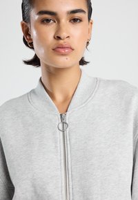 Sweat-shirt gris à fermeture éclair avec un col rond, une fermeture métallique avec un tirette circulaire, et un tissu doux et texturé.