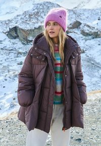Brązowa puchowa kurtka z kapturem i zatrzaskami, noszona na wielobarwnym dzianinowym swetrze. Różowa czapka zimowa i zimowy krajobraz w tle.