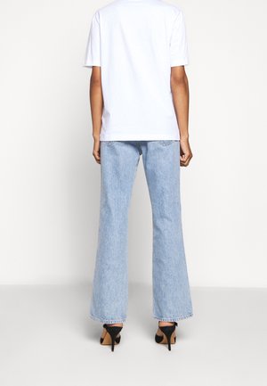 Osoba ubrana w białą koszulę z krótkim rękawem, jasnoniebieskie rozszerzane dżinsy i czarne buty na wysokim obcasie, stojąca tyłem na jednolitym tle.