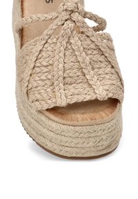 Sandalia de plataforma tejida en color beige, con correas trenzadas y punta abierta, que cuenta con una suela texturizada y una plantilla acolchada.