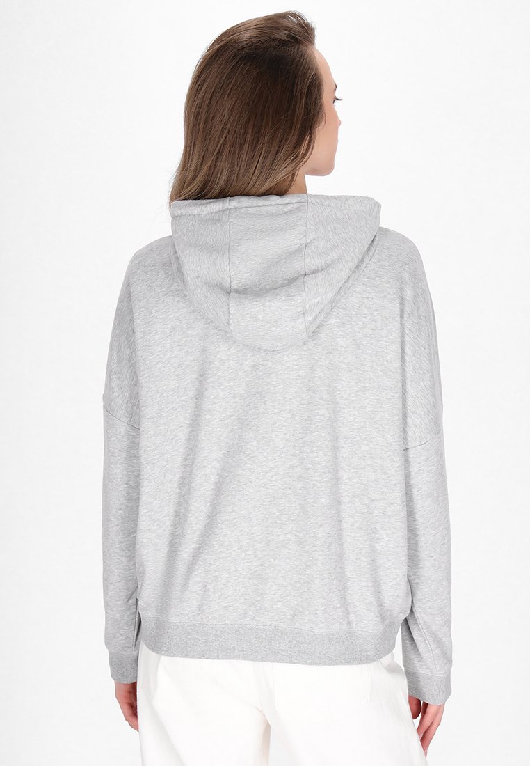 Frau mit langen braunen Haaren, die einen hellgrauen Hoodie und weiße Hosen trägt, mit dem Rücken zu einem schlichten weißen Hintergrund.