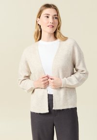 Beige stickad cardigan med V-ringning, öppen front, långa ärmar och fluffig textur; bärs över en vit t-shirt och mörkgrå byxor.