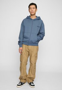 Modrá mikina na zip s volným střihem, kombinovaná s béžovými cargo kalhotami s několika kapsami. Outfit doplňují černobílé tenisky.