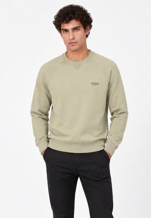 Homme aux cheveux bouclés portant un sweat-shirt beige avec le logo "GUESS" et un pantalon noir, debout les mains dans les poches sur fond blanc.