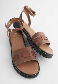 Brune sandaler lavet af læder og ruskind. Har brede stropper, en rem omkring anklen med spænde og en tyk sort sål med hvid stikning.