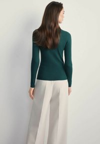 Kobieta z długimi brązowymi włosami, ubrana w dopasowany ciemnozielony sweter prążkowany i szerokie jasnobeżowe spodnie, stojąca tyłem na jednolitym tle.