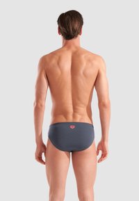 Uomo adulto visto da dietro che indossa slip da bagno grigio scuro con un piccolo logo rosso su uno sfondo grigio chiaro uniforme.