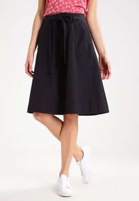 Sort bomulds A-linje nederdel med båndtalje, to forlommer og en glat tekstur, parret med hvide sneakers.
