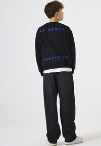 Sudadera negra con texto azul en la parte posterior: "ESTÉ PREPARADO" y "DESAFÍO." Llevada sobre una camiseta blanca, combinada con pantalones negros holgados y zapatillas blancas.