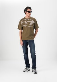 Olivově zelené tričko s krátkým rukávem a bílým grafickým designem, spárováno s tmavě modrými džíny a černými slunečními brýlemi. Doplňují to černé a stříbrné tenisky.