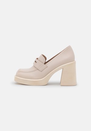 Beige high-heeled shoe with a round toe, slip-on design, decorative strap, and ribbed block heel. Smooth leather texture.