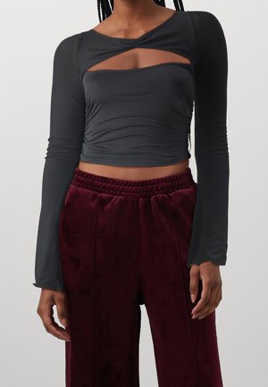 Žena nosící černý crop top s dlouhým rukávem a výstřihem na hrudi a vysoce pasované bordó sametové kalhoty na jednobarevném pozadí.