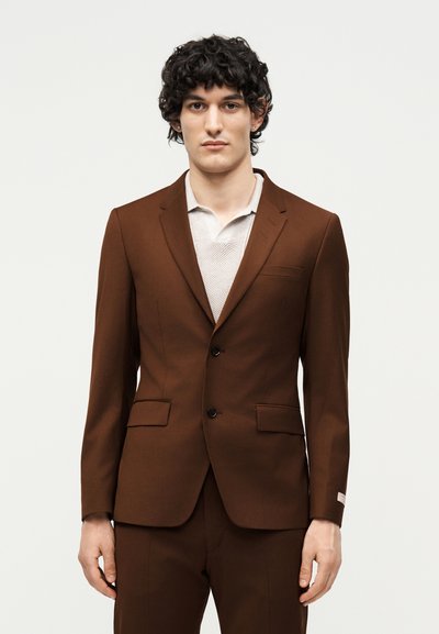 Νέος άνδρας με σγουρά μαλλιά φοράει καφέ σακάκι με δύο κουμπιά πάνω από μια υφασμένη λευκή πόλο, στέκεται έναντι ενός απλού ανοιχτόχρωμου φόντου.