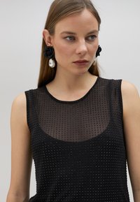 Svart ärmlös topp med en strukturerad mesh-design, prydd med små nitar och en rundad halsringning. Stora blommformade örhängen med kristallhäng.