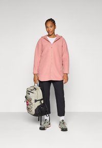 Eine Person steht lächelnd da, trägt eine pinke Fleecejacke mit Reißverschluss, schwarze Hosen und sportliche Schuhe und hält einen hellen Rucksack.