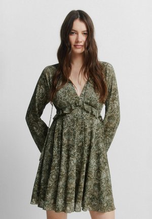 Kvinde med langt brunt hår iført en grøn kjole med paisley-mønster og lange ærmer, stående foran en ensfarvet lys baggrund.