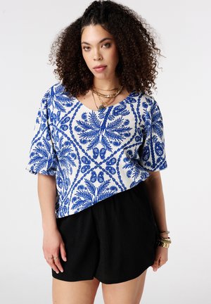 Vrouw met krullend haar die een blauw-witte blouse met patroon en zwarte korte broek draagt, geaccentueerd met gelaagde kettingen en armbanden.