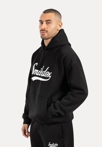 Sudadera negra de algodón, con un bolsillo canguro, un logo blanco en el pecho y puños y dobladillo acanalados. Diseño simple, sin adornos.