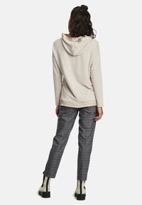 Kvinna med mörkt hår i en krämfärgad huvtröja, grå rutiga byxor och vita stövlar med svarta ränder som står med ryggen mot kameran mot vit bakgrund.