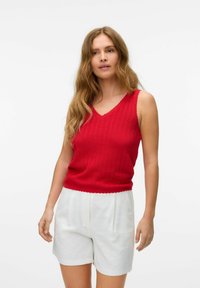 Rød strikket tanktop med v-hals og bølgede kanter, parret med hvide plisserede shorts. Materialet er en bomuldsblanding med en struktureret overflade.