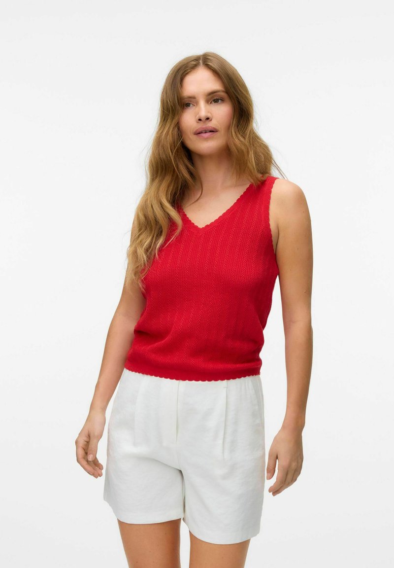 Rød strikket tanktop med v-hals og bølgede kanter, parret med hvide plisserede shorts. Materialet er en bomuldsblanding med en struktureret overflade.