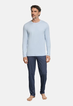 Uomo che indossa una camicia a maniche lunghe azzurro chiaro e pantaloni del pigiama blu scuro con motivi, in piedi a piedi nudi, che guarda leggermente di lato.