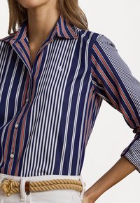 Camicia a righe navy con linee bianche e rosse, chiusura con bottoni e colletto. Presenta una leggera lucentezza con un logo sottile sul petto.