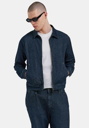 Giacca in denim blu scuro con zip frontale, colletto e tasche laterali, indossata sopra una t-shirt bianca. Texture liscia con dettagli di cucitura.