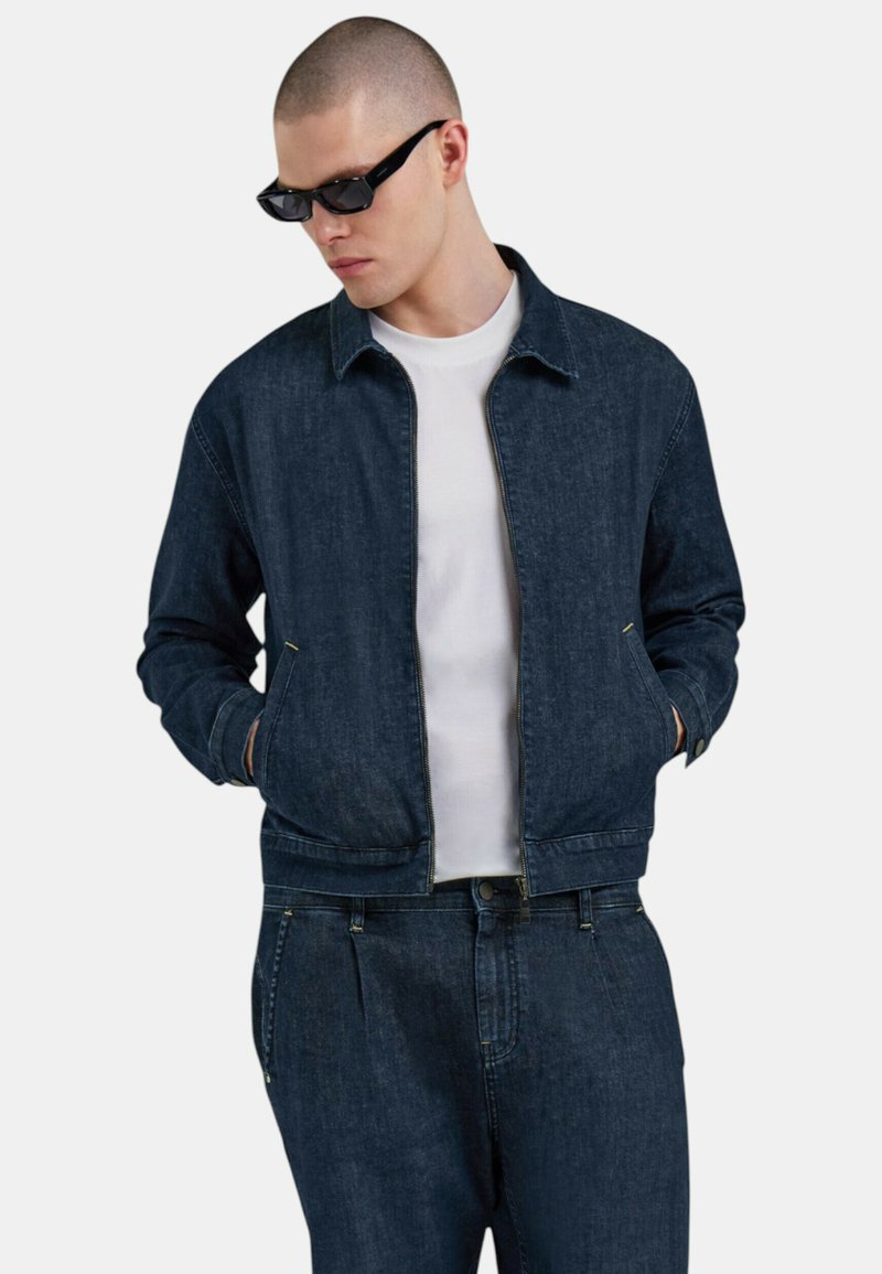 Giacca in denim blu scuro con zip frontale, colletto e tasche laterali, indossata sopra una t-shirt bianca. Texture liscia con dettagli di cucitura.