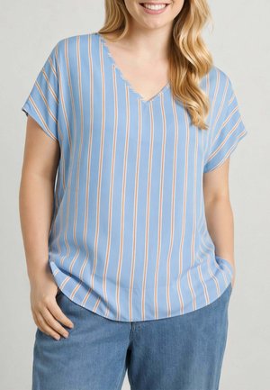 Donna che indossa una maglietta blu a maniche corte con scollo a V, righe verticali bianche e arancioni e jeans blu, sorridente con le mani in tasca.