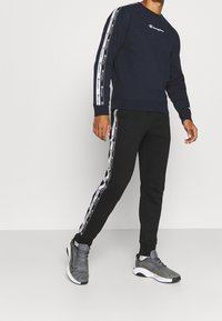Sudadera de color navy con logo blanco y franjas laterales, combinada con joggers negros que presentan franjas laterales a juego, y zapatos deportivos grises.