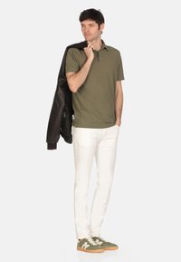 Polo shirt verde oliva, pantaloni slim-fit bianchi, sneakers grigio-verdi con suola chiara. Indossa una giacca di pelle nera appoggiata su una spalla.